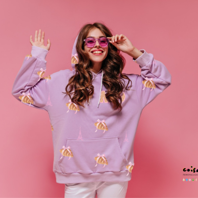 Mulher sorridente com sweatshirt lilás estampado de abelhas amarelas e torres Eiffel, usando óculos sol rosa, fundo rosa