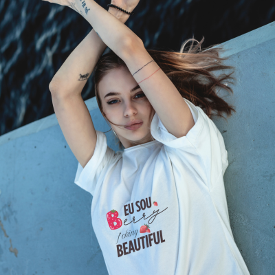 Modelo jovem com t-shirt branca estampada, posando junto a uma parede cinza e um corpo de água