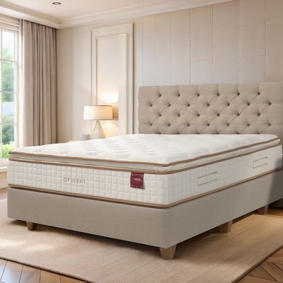 Cama com colchão bege claro e cabeceira estofada numa sala iluminada com duas mesas de cabeceira e plantas.