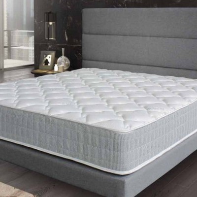 Colchão branco acolchoado sobre base cinza com cabeceira, numa sala com chão em madeira escura e janelas grandes