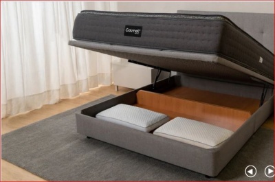 Cama cinzenta com colchão levantado e espaço de arrumação com almofadas duas almofadas