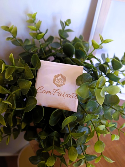 Pacote branco com texto ComPaixão rodeado de folhas verdes artificiais