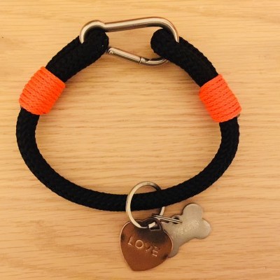 Pulseira preta de corda com detalhes laranja e pendentes metálicos em fundo de madeira clara