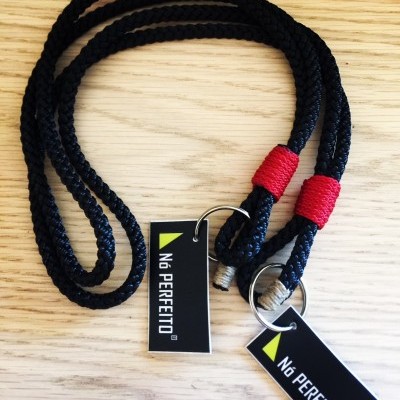 Cordão preto trançado com acabamentos vermelhos e etiquetas NÓ PERFEITO sobre madeira clara