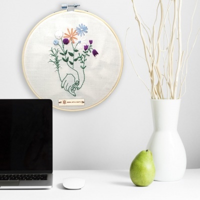 Quadro bordado redondo com flores e mãos entrelaçadas na parede sobre mesa com computador, vaso e pera