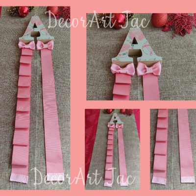 Peça decorativa letra A em madeira com laços e fitas cor de rosa sobre tecido bege