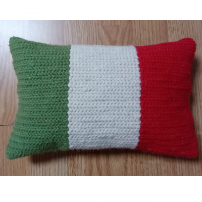 Travesseiro rectangular com capa tricotada nas cores verde, branco e vermelho sobre piso de madeira.