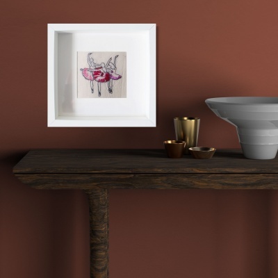 Moldura branca com bordado floral em tecido bege numa parede castanha, mesa de madeira escura com copos dourados e vaso cinza.