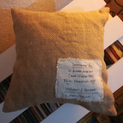 Almofada de tecido bege com etiqueta manuscrita costurada, em ambiente com livros e tapete.