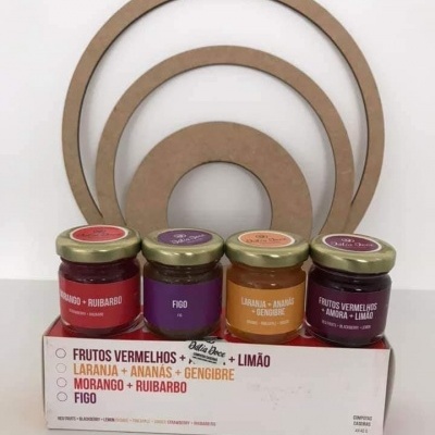 Quatro frascos de compotas de frutas com etiquetas coloridas numa caixa branca com texto e círculos de madeira ao fundo