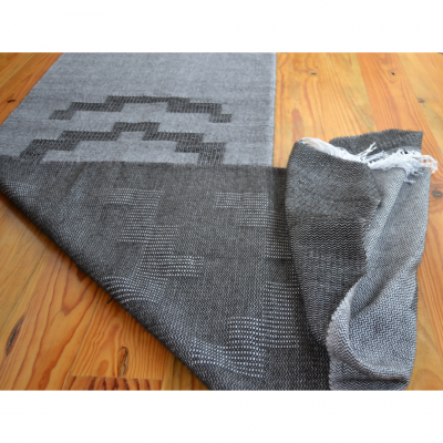 Tapete cinza com padrão geométrico sobre chão de madeira clara, com ponta dobrada mostrando o avesso.