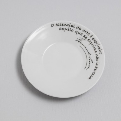 Prato branco redondo com frase em português no bordo e assinatura, fundo cinza