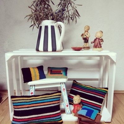 Conjunto artesanal com almofadas coloridas, jarro com riscas pretas e figuras em cerâmica sobre mobiliário branco em madeira.