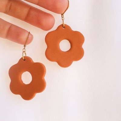 Brincos castanhos em forma de flor com buraco central e gancho dourado sendo segurados por uma mão