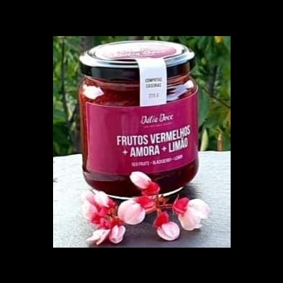 Frasco de geleia Dália Doce com frutas vermelhas, amora e limão, sobre superfície clara com flores rosas