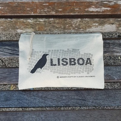 Necessaire branca com padrão de azulejos cinzentos, pássaro preto e texto LISBOA colocada num banco de madeira.