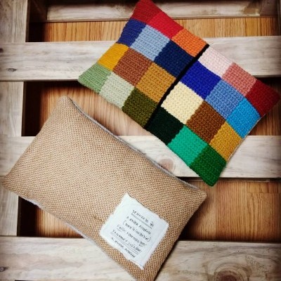 Três almofadas, duas coloridas em crochê e uma bege com etiqueta, sobre superfície de madeira clara