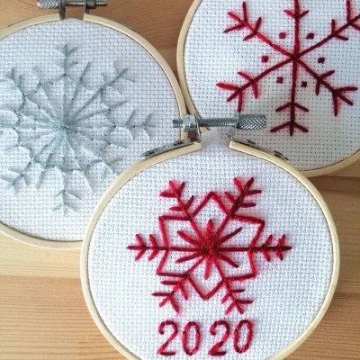 Três bastidores de bordado com flocos de neve bordados em branco, vermelho e cinza, um com texto 2020.