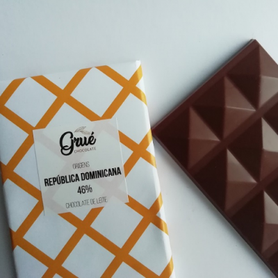 Barra de chocolate ao leite da marca Grué com embalagem branca e laranja e etiqueta com texto, ao lado da barra de chocolate marrom com padrão em relevo