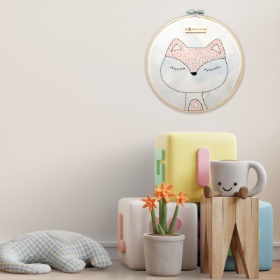 Cenário infantil com bordado de raposa, blocos de brinquedo coloridos, copo com sorriso, almofada xadrez e vaso com flores.