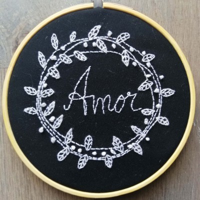 Bastidor com bordado branco em tecido preto com a palavra 'Amor' rodeada por folhas