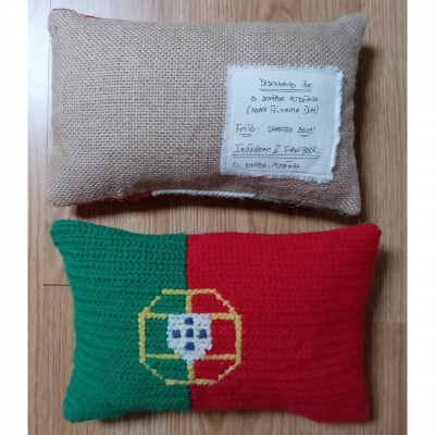 Duas almofadas retangulares sobre chão de madeira clara, uma em juta bege com etiqueta branca manuscrita e outra em crochê na bandeira de Portugal.