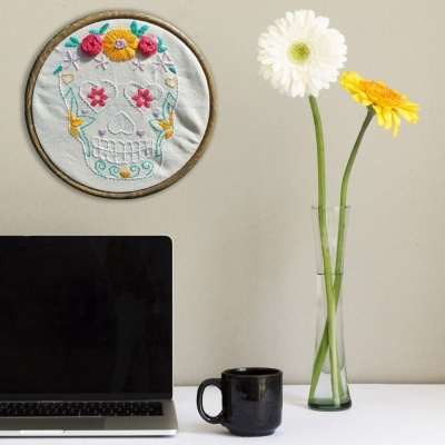 Bordado de caveira colorida, portátil, caneca preta e vaso com flores numa mesa
