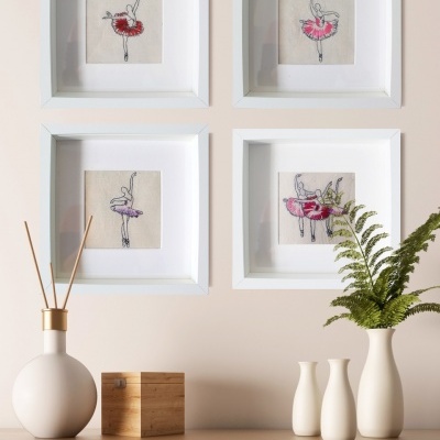 Quadros de bailarinas bordadas em saias vermelha e rosa na parede cima de móvel com vasos e caixa.