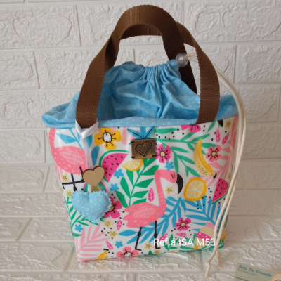 Bolsa colorida com padrão de flamingos, frutas e folhas e alças castanhas, sobre fundo branco texturado