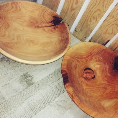 Dois pratos grandes de madeira com padrões naturais e imperfeições, sobre chão de madeira clara e junto a parede de tábuas.