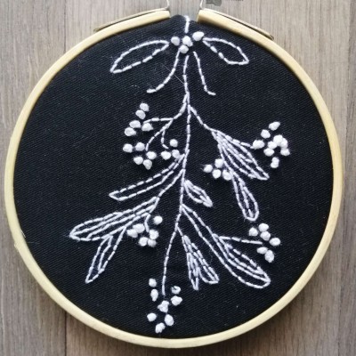 Aro de bordado com tecido preto e bordado branco de ramo com folhas e bagas, sobre fundo de madeira cinzenta