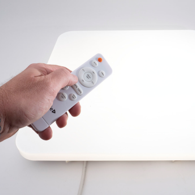 Controle remoto branco segurado por uma mão em frente a uma luminária retangular branca iluminada