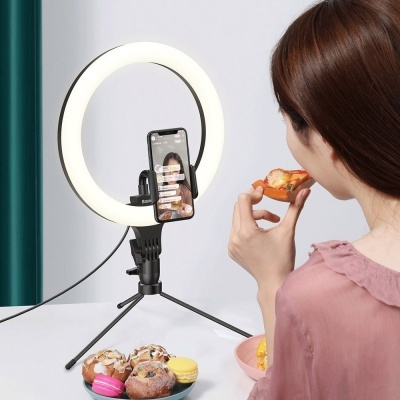 Anel de luz LED branco com suporte para telemóvel e tripé preto em mesa com mulher a comer pizza e pratos com pastelaria