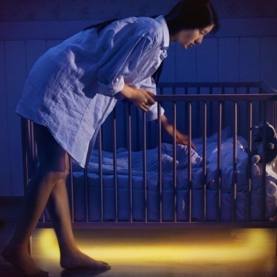 Berço de madeira com iluminação amarela sob ele e mulher inclinada para dentro do berço.