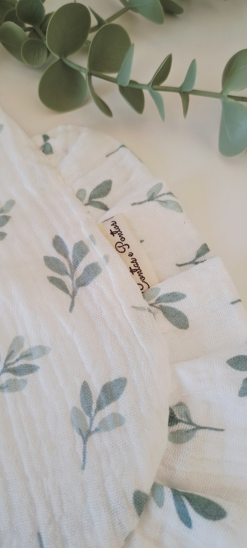 Babete ♥ eucalyptus leaf Pano branco com padrão de folhas verdes e etiqueta Quintal do Sonhador em superfície branca