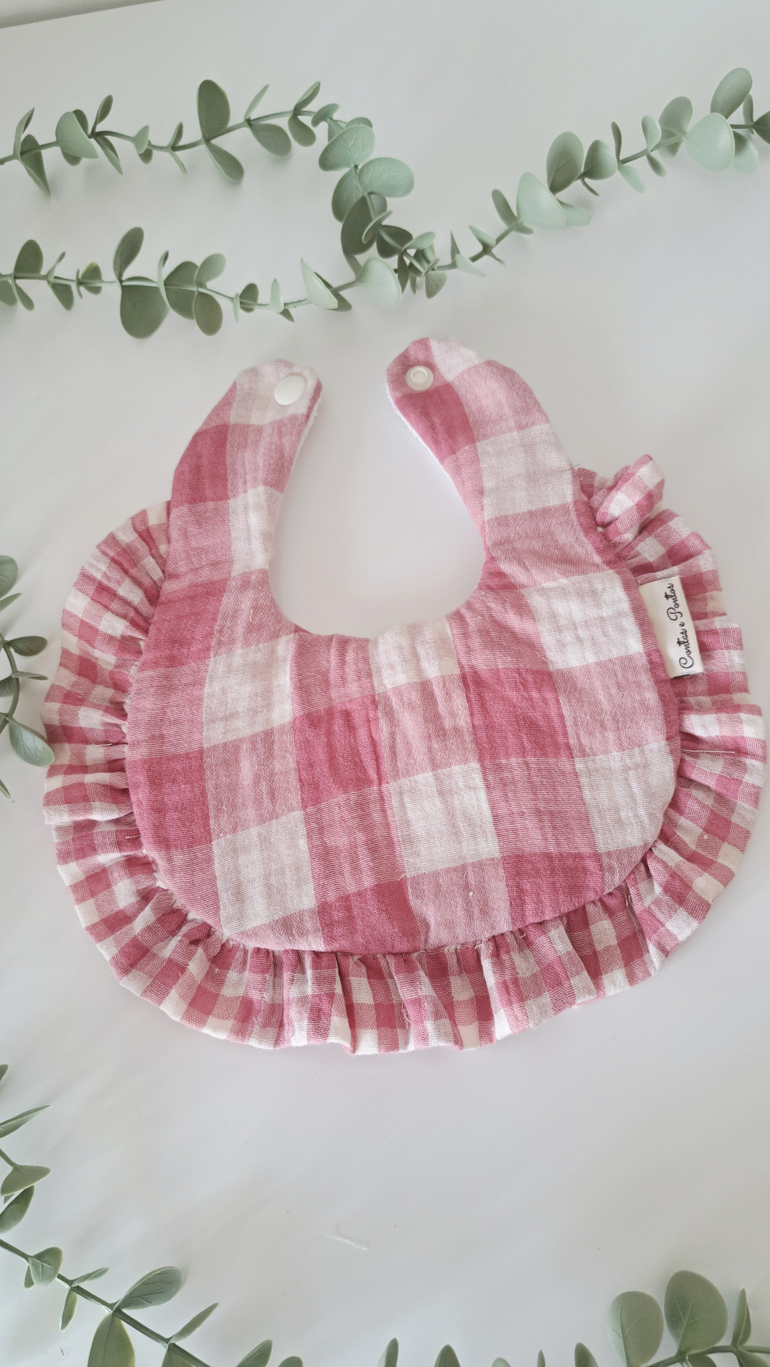 Babete ♥ large vichy - rose Babete para bebé com padrão xadrez rosa e branco, com bordas plissadas, colocado sobre uma mesa branca cercada por folhagem verde.