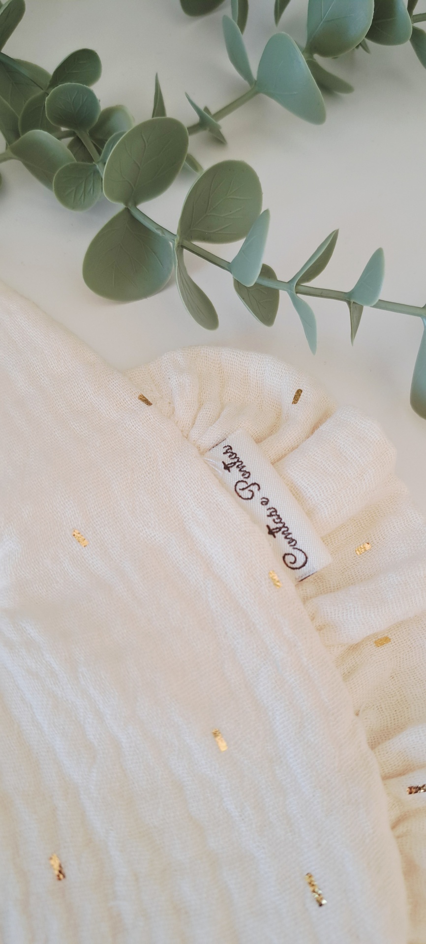 Babete sand golden foil tecido branco com detalhes dourados e etiqueta com texto, ramos verdes artificiais em fundo branco