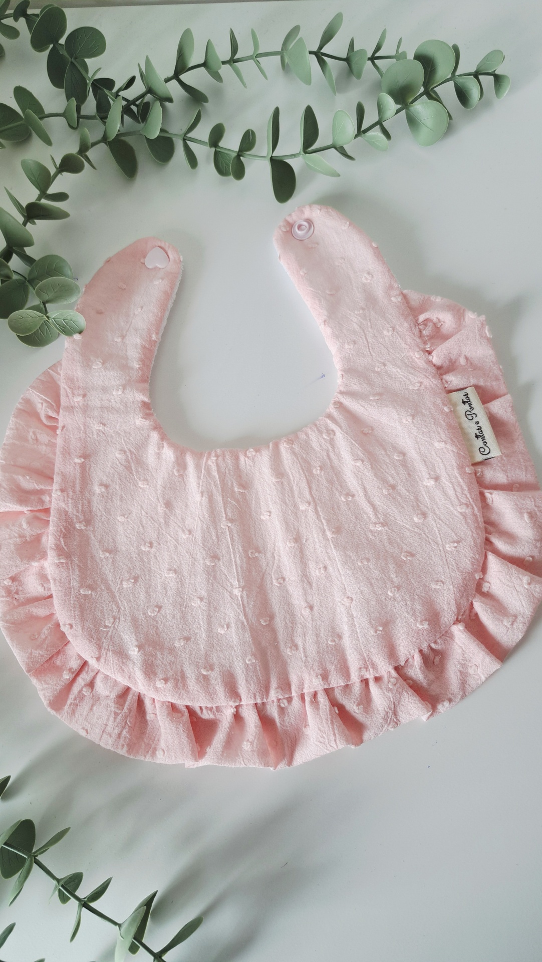 Babete ♥ pale rose Babete cor-de-rosa claro com textura, ruffles, botão de pressão e etiqueta, sobre superfície branca com ramos verdes
