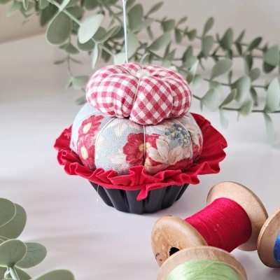 Alfineteiro em tecido com padrões xadrez e floral, base preta com babado vermelho, rolos de linha colorida e folhas verdes ao redor