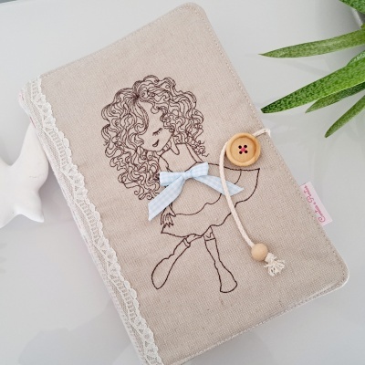 Capa de tecido bege com bordado de menina, renda branca e botão de madeira