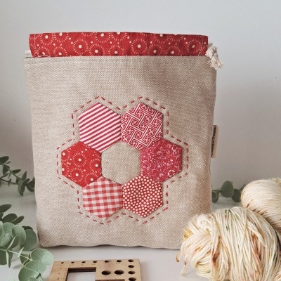 Bolsa de tecido bege com padrões hexagonais vermelhos e brancos, bola de lã, régua de madeira e ganchos metálicos