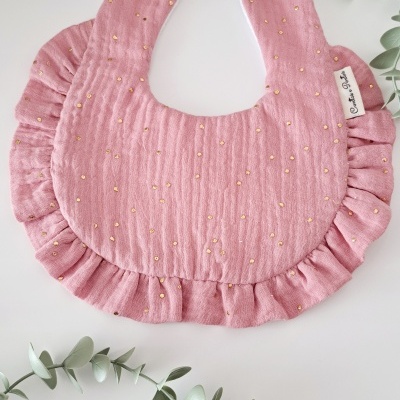 Babete de bebé cor-de-rosa com pontinhos dourados e volantes, com fecho de pressão, em fundo branco com folhas verdes