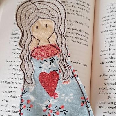Marcador de livros artesanal em tecido com desenho de boneca pendurado num livro aberto