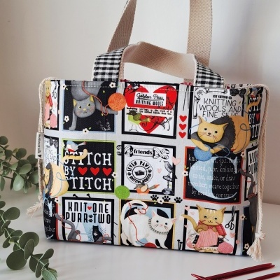 Bolsa de tecido decorada com desenhos de gatos e novelos de lã, com alças quadriculadas pretas e brancas.