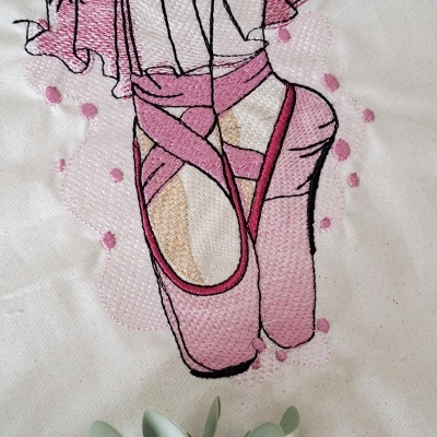 Bordado de pés de bailarina com sapatilhas cor de rosa em tecido branco, com ramo de folhas verdes abaixo.