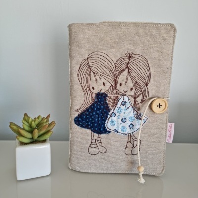 Capa bege com desenho bordado de duas meninas e botão de fecho, ao lado de planta suculenta em vaso branco.
