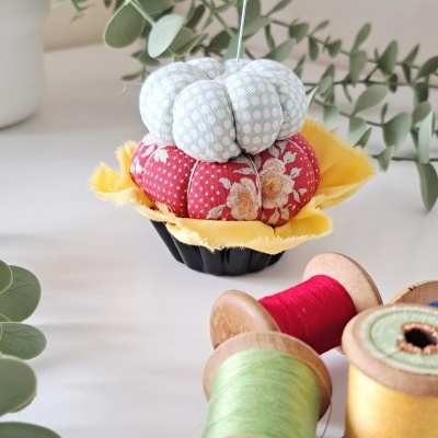 Alfineteiro em tecido com carretéis de linha ao lado e plantas verdes no fundo.