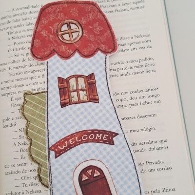 Marcador de livros em tecido com design de casa em fundo branco com folhas verdes ao fundo