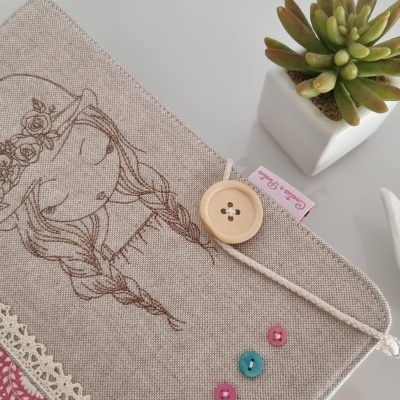 Capa de tecido bege bordada com menina e detalhes em renda, botões coloridos, numa mesa branca com planta suculenta e figura de pássaro.