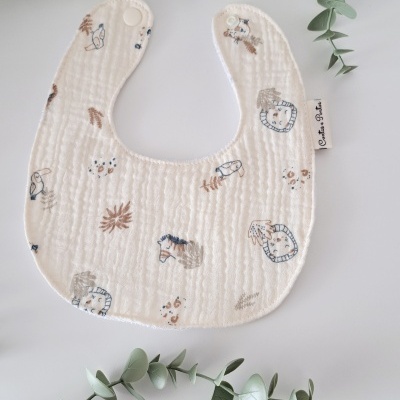 Babete branco com estampado de animais e folhas e fecho de pressão, rodeado por folhas verdes